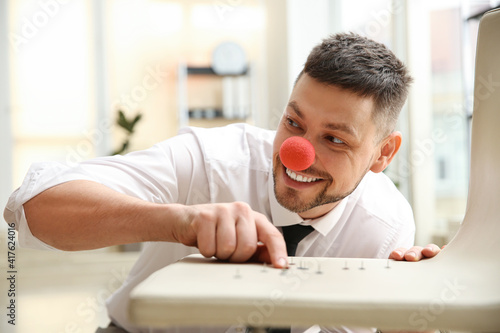 Man with clown nose putting pins on colleague's chair in office. Funny joke photo