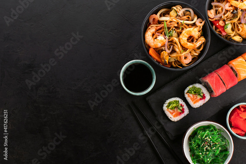 Assorted sushi rolls and wok noodles with shrimps and vegetables in a black bowl, seaweed salad on a black slate background, top view, copy space. Traditional Asian food set.
