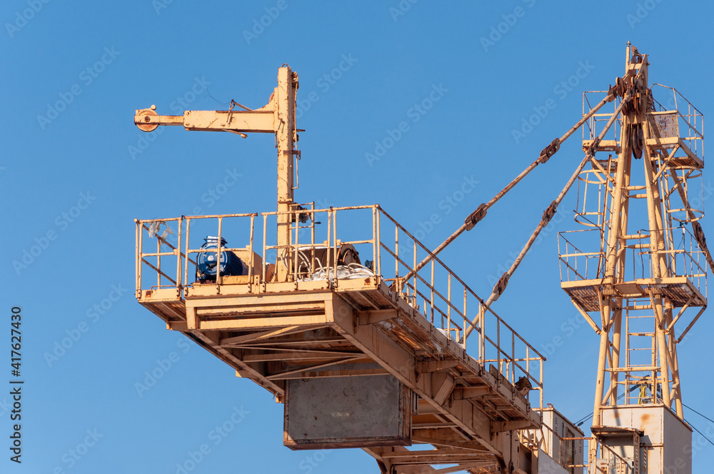 Fragment of a tower crane in close-up against a blue clear sky. Yellow painted metal structure, main winch, counterweight, tower peak, rear pendant.