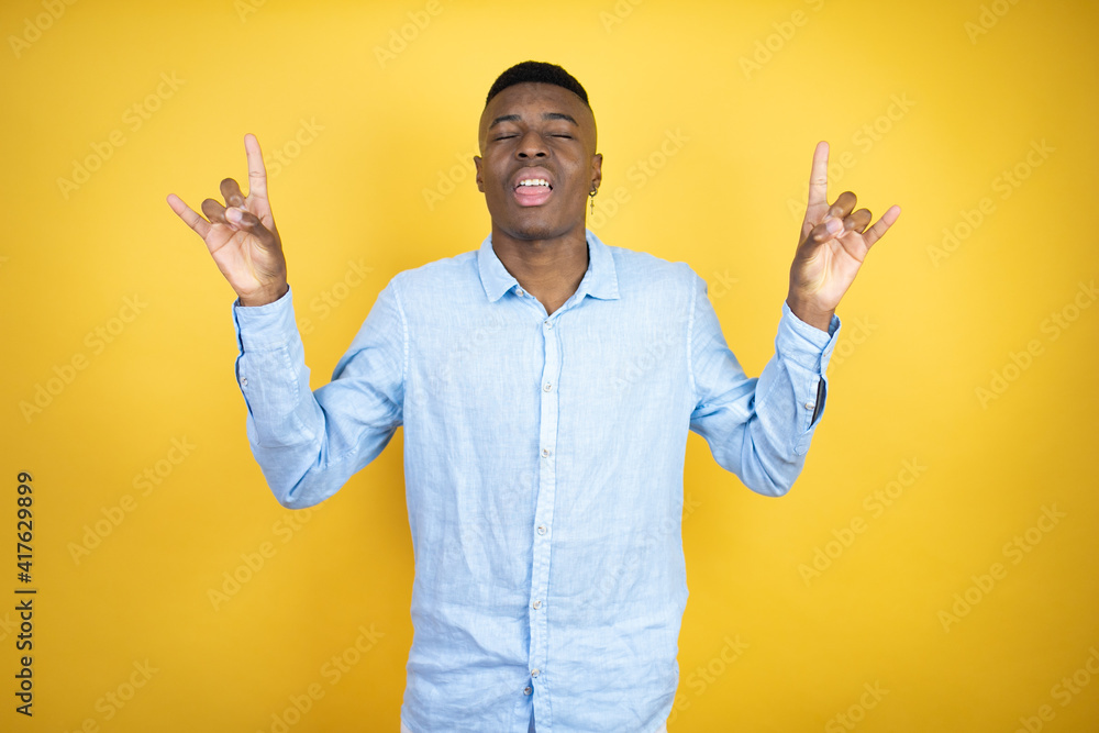 Young african american man wearing a casual shirt standing over yellow background shouting with crazy expression doing rock symbol with hands up Young african american man wearing a casual shirt standing over yellow background shouting with crazy expression doing rock symbol with hands up
