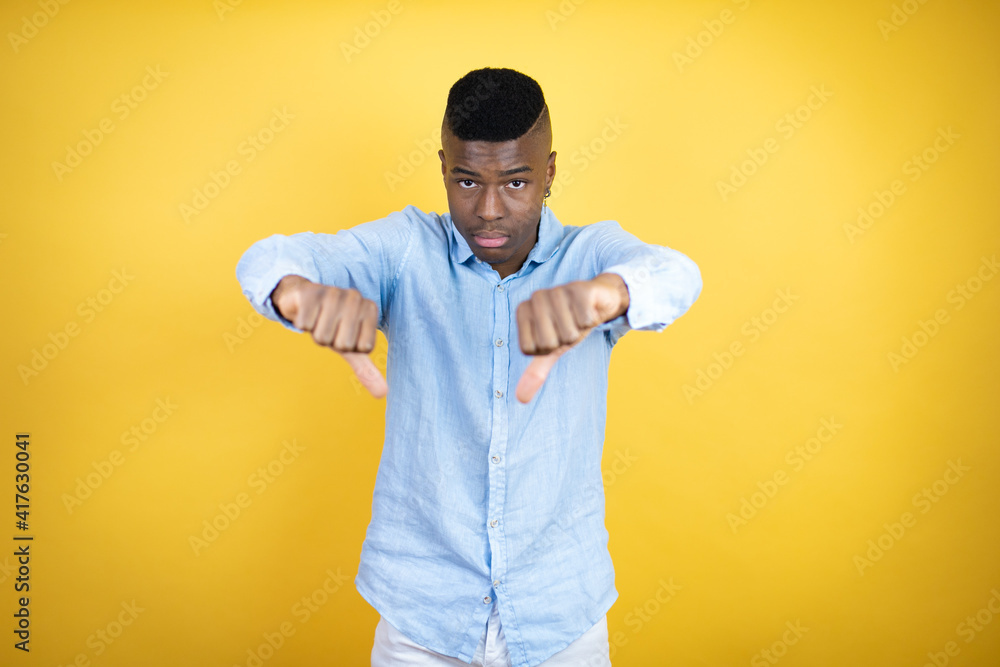 Young african american man wearing a casual shirt standing over yellow background with angry face, negative sign showing dislike with thumbs down Young african american man wearing a casual shirt standing over yellow background with angry face, negative sign showing dislike with thumbs down