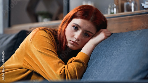 Upset red haired teenager looking at camera on couch