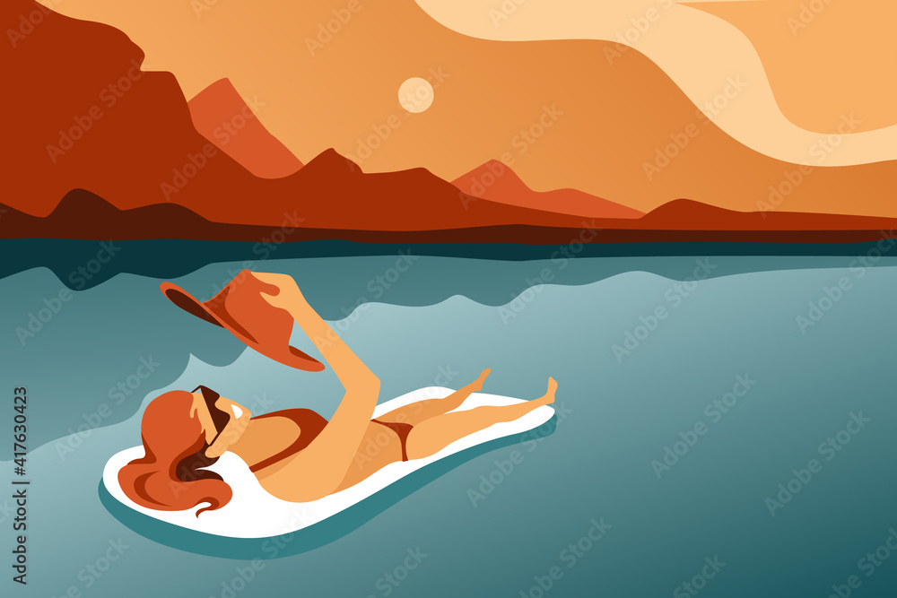 A young girl in a bathing suit and sun glasses bathes on a mattress in the sea against the background of abstract mountains and sky. The girl sunbathes on an inflatable mattress, a hat in her hands.