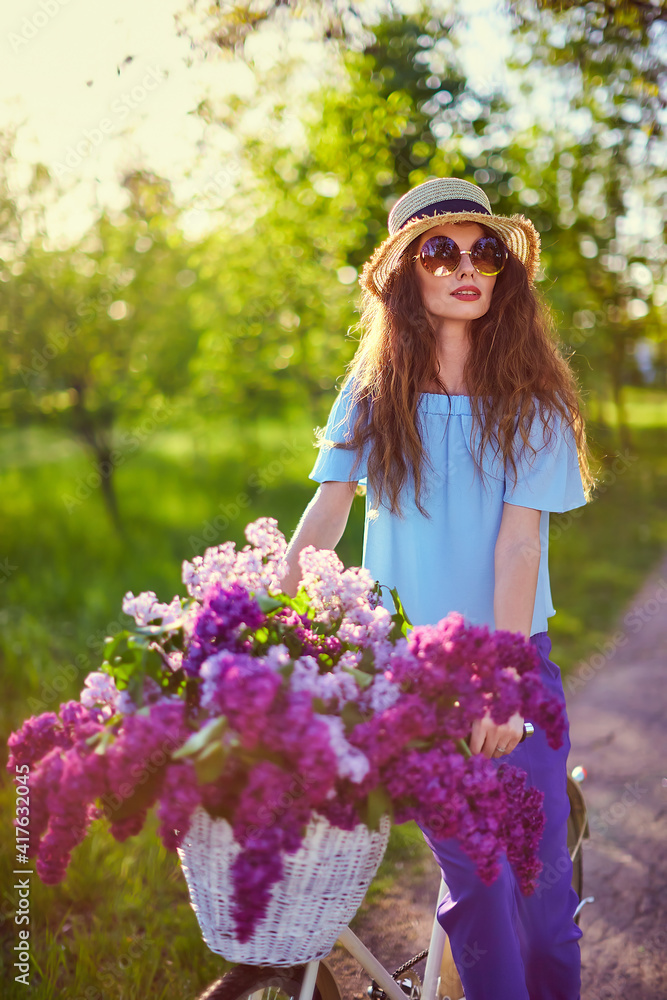 Portrait of a happy beautiful woman in straw hat with vintage bicycle and flowers on park background in the sunlight outdoor. Bike with basket full of flowers. Active Leisure Concept. Portrait of a happy beautiful woman in straw hat with vintage bicycle and flowers on park background in the sunlight outdoor. Bike with basket full of flowers. Active Leisure Concept.