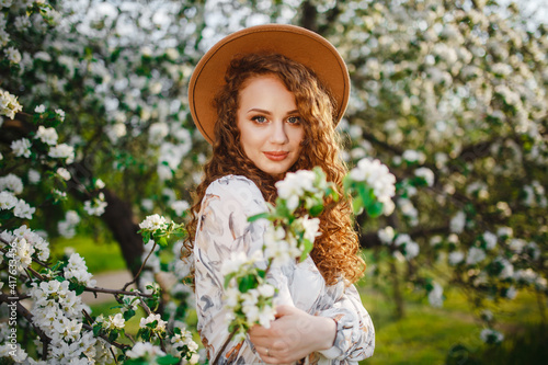 Portrait shot of a young girl near the blooming white tree at the spring park. Female wearing beige hat and white dress standing among blossom on the apple tree. Spring season concept.