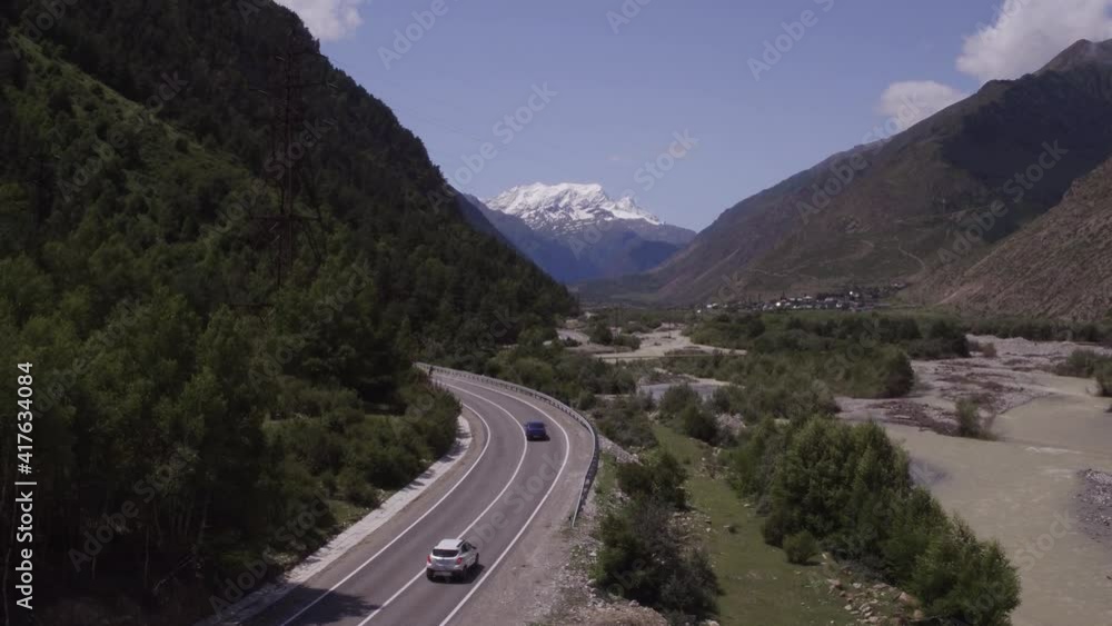 Two car driving on asphalt serpentine road through gorge in mountains with river along. Aerial drone wide shot at summer cloudy day Two car driving on asphalt serpentine road through gorge in mountains with river along. Aerial drone wide shot at summer cloudy day