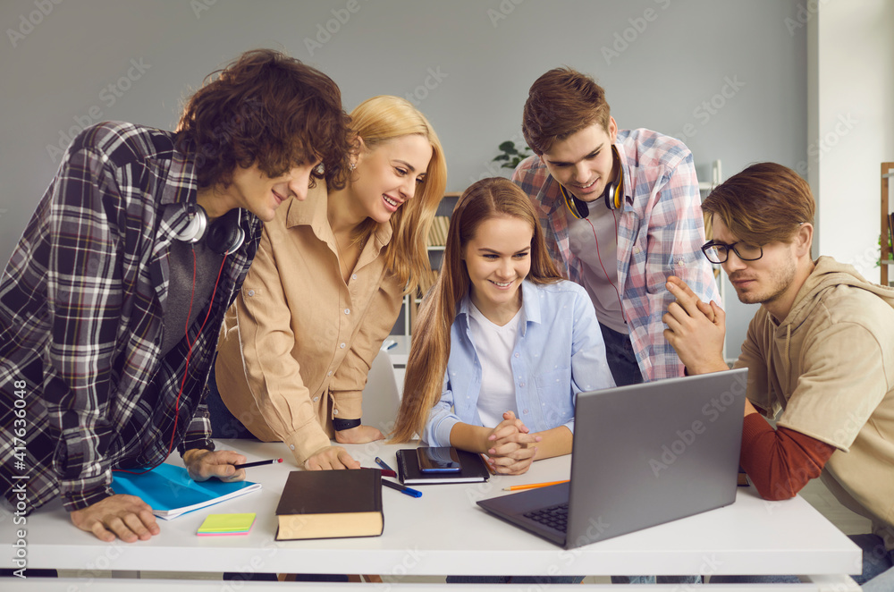 Group of happy male and female students looking at screen of modern laptop computer. School, university or college mates working on digital project together. Secondary and higher education concept