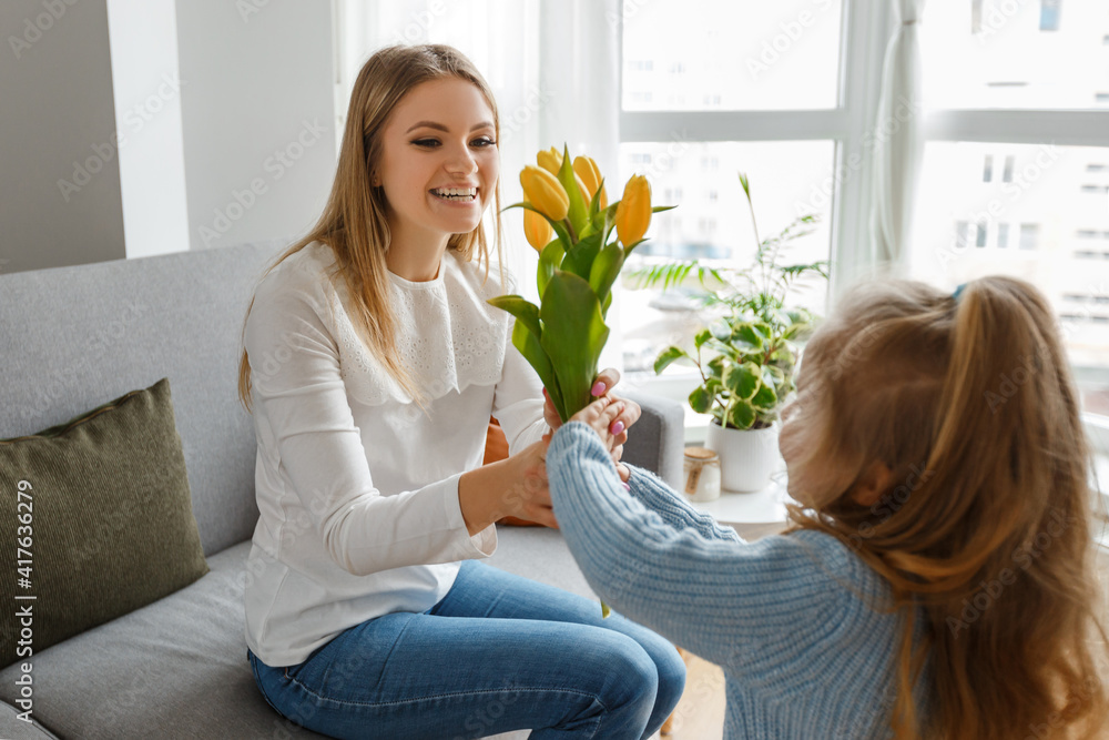 International women's and mother's day. Little cute daughter with bouquet of yellow flowers congratulates her smiling mom. Happy family holidays at home. Curly girl surprise mommy International women's and mother's day. Little cute daughter with bouquet of yellow flowers congratulates her smiling mom. Happy family holidays at home. Curly girl surprise mommy