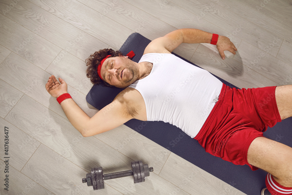 Funny tired fat man lying on fitness mat after gym workout. Exhausted guy  in retro sweatband,
