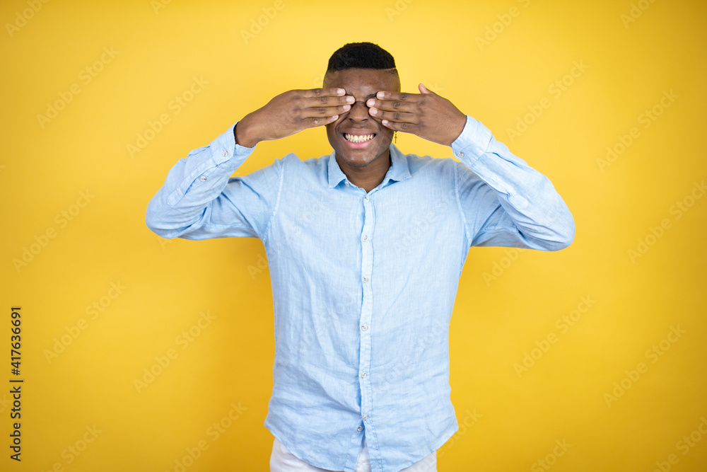 Young african american man wearing a casual shirt standing over yellow background covering eyes with hands smiling cheerful and funny. Blind concept. Young african american man wearing a casual shirt standing over yellow background covering eyes with hands smiling cheerful and funny. Blind concept.