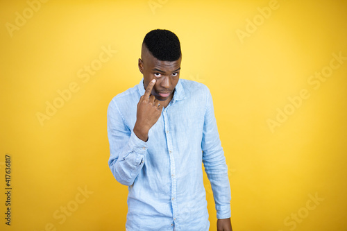 Young african american man wearing a casual shirt standing over yellow background Pointing to the eye watching you gesture, suspicious expression