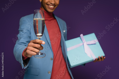 partial view of cheerful african american man holding glass of champagne and present on purple