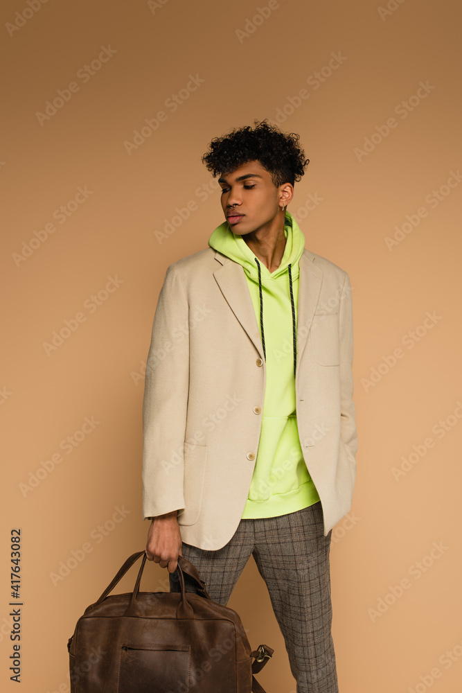Fototapeta premium trendy african american man in hoodie and blazer holding leather bag on beige