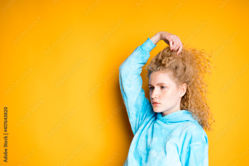 A young girl with her hand straightens her blond hair while standing on a bright background A young girl with her hand straightens her blond hair while standing on a bright background