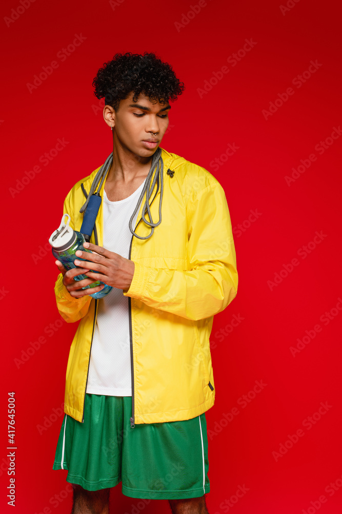 curly african american man in yellow rain jacket standing with skipping rope and sports bottle isolated on red curly african american man in yellow rain jacket standing with skipping rope and sports bottle isolated on red