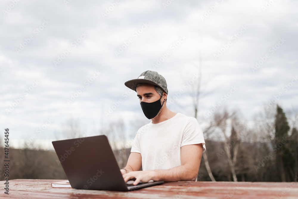 Man wearing mask and cap working with a laptop on a table outdoors