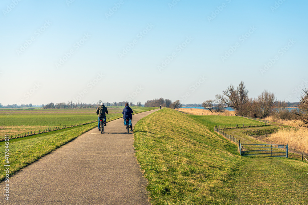 An unidentified man and woman are cycling on a winding Dutch dike near the village of Drimmelen in the province of Noord-Brabant. It's a sunny day at the end of the winter season.