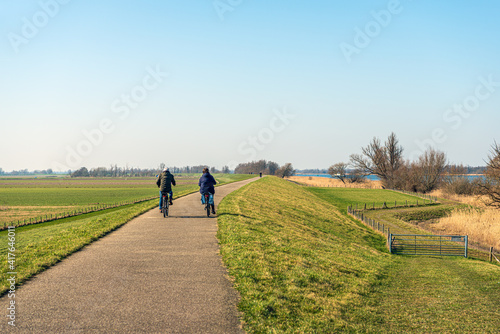 An unidentified man and woman are cycling on a winding Dutch dike near the village of Drimmelen in the province of Noord-Brabant. It's a sunny day at the end of the winter season.