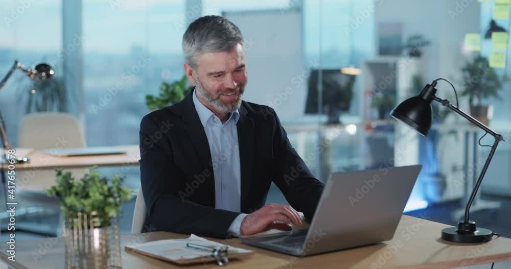 Cheerful business manager working on portable laptop sitting at desk. Mature adult businessman inside modern office interior. Video conference. Emotions. Cheerful business manager working on portable laptop sitting at desk. Mature adult businessman inside modern office interior. Video conference. Emotions.