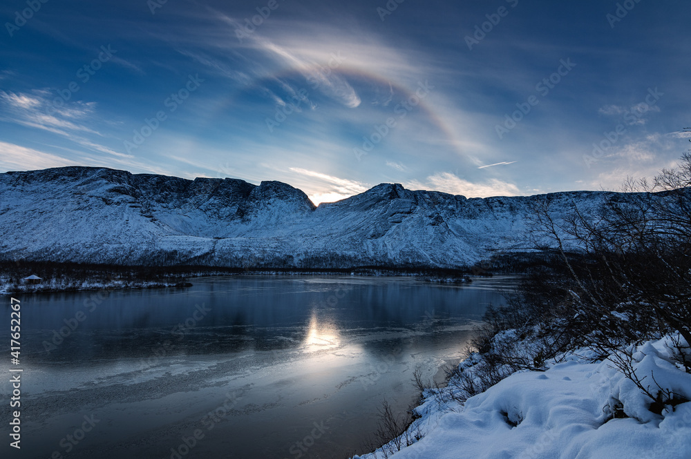 Solar halo in the mountains near a lake covered with ice on a winter evening