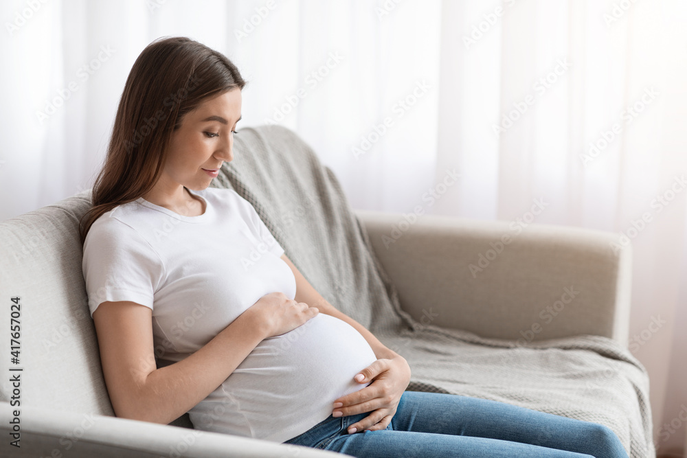 Prenatal Time. Beautiful Future Mother Sitting On Couch Embracing Her Belly Prenatal Time. Beautiful Future Mother Sitting On Couch Embracing Her Belly
