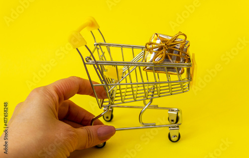 Female hand touching mini shopping trolley with gift inside on yellow background Minimalistic shopping concept