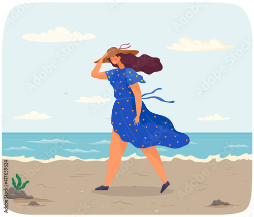 Young woman walking alone on beach. Lady hold her hat on head because of windy weather. Person dressed in blue dotted dress. Beautiful landscape of ocean bank. Girl is walking during strong wind