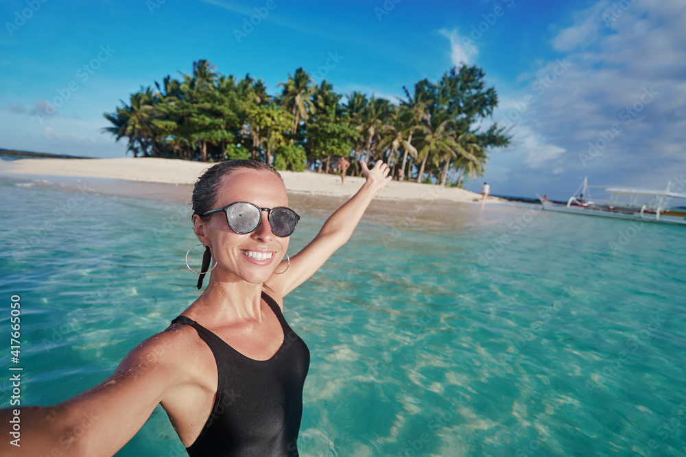 Traveling in Philippines. Happy young woman taking selfie while bathing near small island with white sand tropical beach.