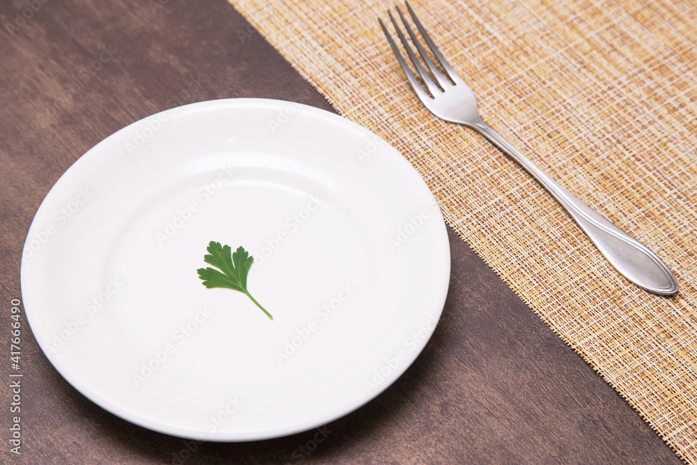 Single one parsley green leaf on white empty plate and fork on table. Diet, fasting, vegan, vegetarian, healthy food concept Single one parsley green leaf on white empty plate and fork on table. Diet, fasting, vegan, vegetarian, healthy food concept
