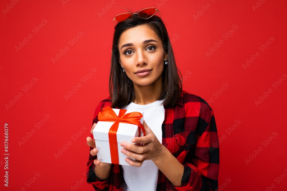 Photo shot of pretty positive young brunette woman isolated over red background wall wearing white casual t-shirt and red and black shirt holding white gift box with red ribbon and looking at camera Photo shot of pretty positive young brunette woman isolated over red background wall wearing white casual t-shirt and red and black shirt holding white gift box with red ribbon and looking at camera