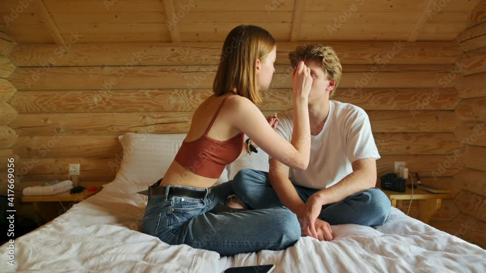 Woman doing makeup to her boyfriend on the bed in the apartment with wooden interior. Woman farbuvaye makeup man on the bed at the hotel. Woman doing makeup to her boyfriend on the bed in the apartment with wooden interior. Woman farbuvaye makeup man on the bed at the hotel.