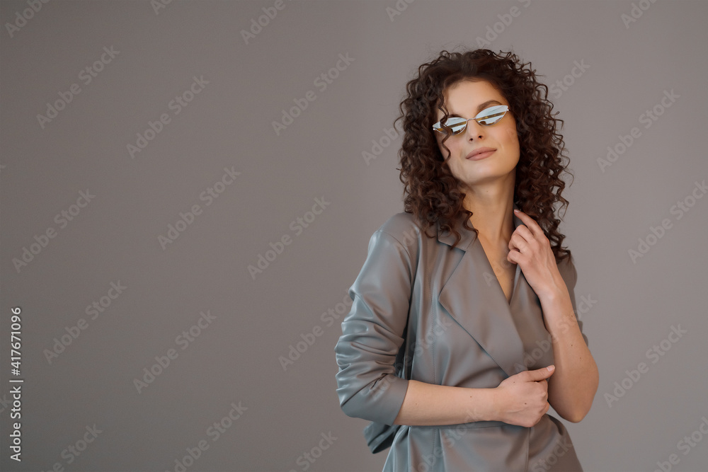 Photo of charming woman 30s with long curly hairstyle wearing trendy sunglasses, leather gray coat, with fashion look over gray background Photo of charming woman 30s with long curly hairstyle wearing trendy sunglasses, leather gray coat, with fashion look over gray background