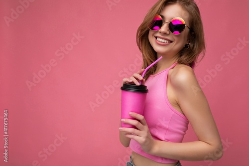 Shot of attractive young happy smiling blonde female person wearing pink top and colourful sunglasses isolated over pink background holding coffee paper cup for cutout drinking and enjoying