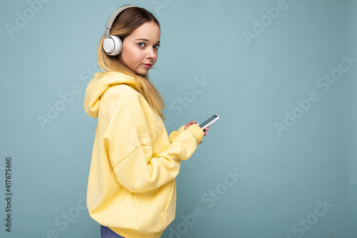 Side-profile beautiful young blonde woman wearing yellow stylish hoodie isolated on blue background wearing white bluetooth wireless headphones and listening to cool music and using mobile phone