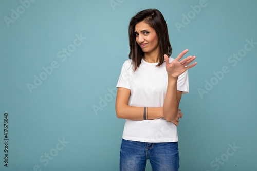 Portrait of dissatisfied emotional young pretty nice winsome brunette woman with sincere emotions wearing casual white t-shirt for mockup isolated over blue background with copy space