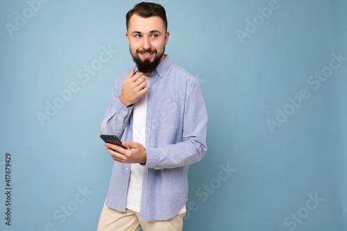 Photo shot of handsome positive good looking young man wearing casual stylish outfit poising isolated on background with empty space holding in hand and using mobile phone messaging sms looking to the