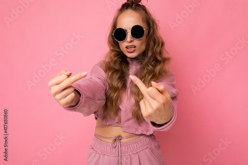 Young self-confident cool beautiful blonde wavy-haired woman with sincere emotions wearing stylish pink sport suit and sunglasses isolated on pink background with copy space and showing middle finger