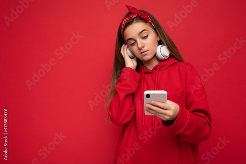 Photo of beautiful concentrated brunette girl wearing red hoodie isolated on red background holding and using smartphone wearing white wireless earphones listening to cool music looking at gadjet photo