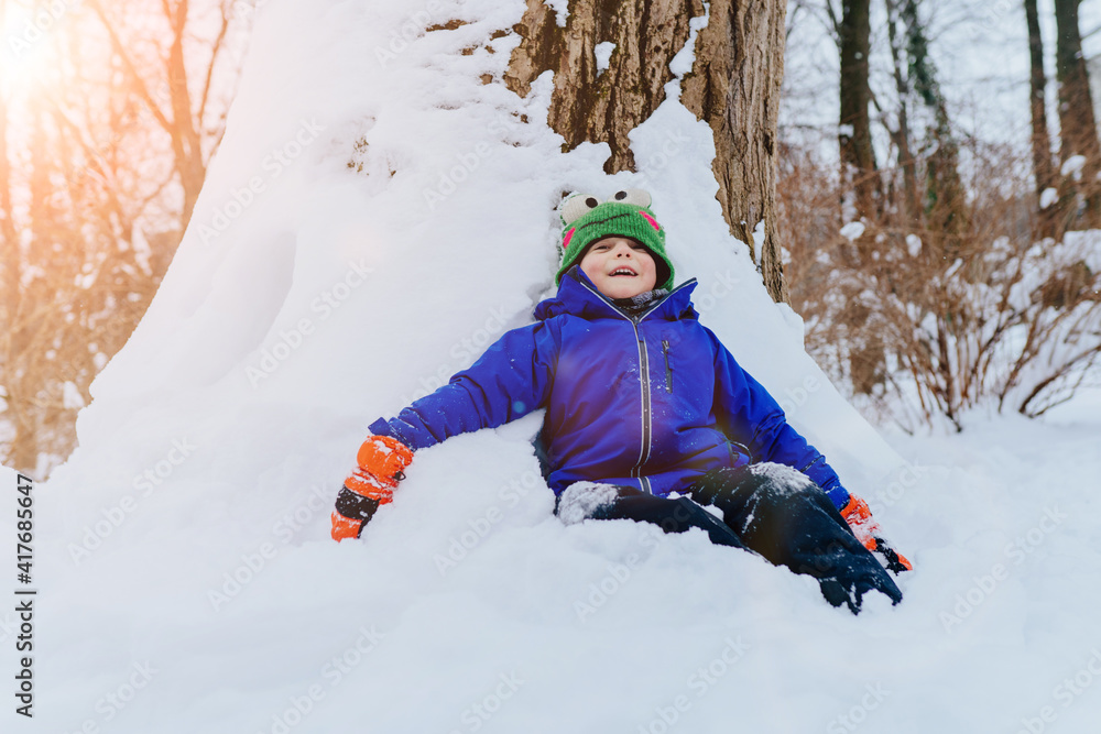 Cute child dressed in a blue jacket, pants, green hat and scarf, gloves, walks through the snowy park enjoying first snow blowing. Sun glare effect. Cute child dressed in a blue jacket, pants, green hat and scarf, gloves, walks through the snowy park enjoying first snow blowing. Sun glare effect.