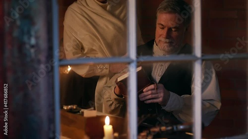 View thru a window of scene from late 1800s of a man sitting at a small desk whose wife enters and brings him his leather bound journal.