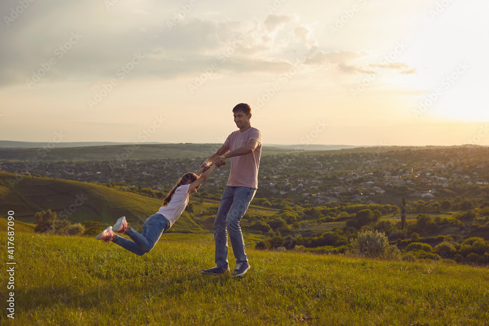 Fototapeta premium Happy fathers day. Young father playing game with his daughter in countryside at sunset, copy space. Dad spinning child in circles outdoors