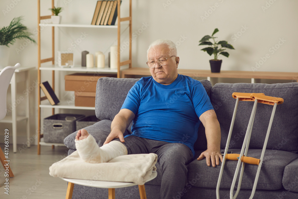 © Studio Romantic - Rehabilitation after accident: Senior patient with bone fracture spends recovery period at home. Sad old man with broken leg in plaster cast sitting on sofa in living-room, thinking about his injury