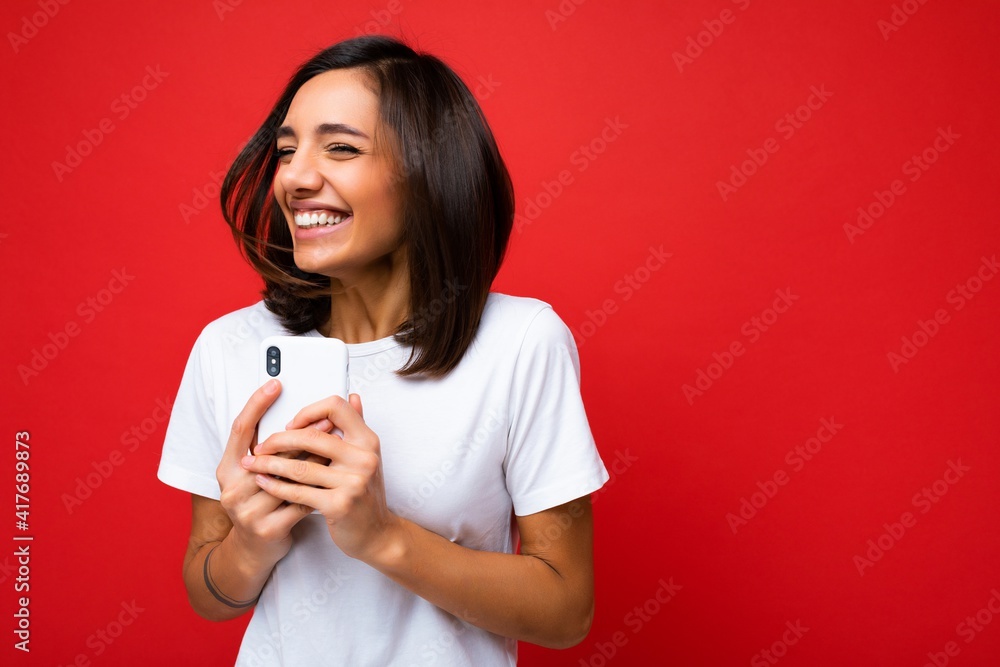 Photo shot of attractive positive happy smiling good looking young brunette woman wearing casual stylishwhite t-shirt poising isolated on red background with empty space holding in hand and using Photo shot of attractive positive happy smiling good looking young brunette woman wearing casual stylishwhite t-shirt poising isolated on red background with empty space holding in hand and using