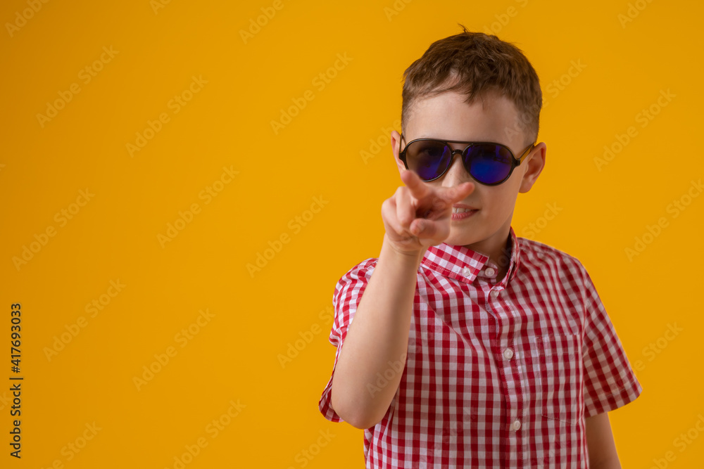 suspicious boy in dark sunglasses and a red checked shirt looks doubtfully at the camera., standing against the yellow background of the studio. Observes the environment suspicious boy in dark sunglasses and a red checked shirt looks doubtfully at the camera., standing against the yellow background of the studio. Observes the environment
