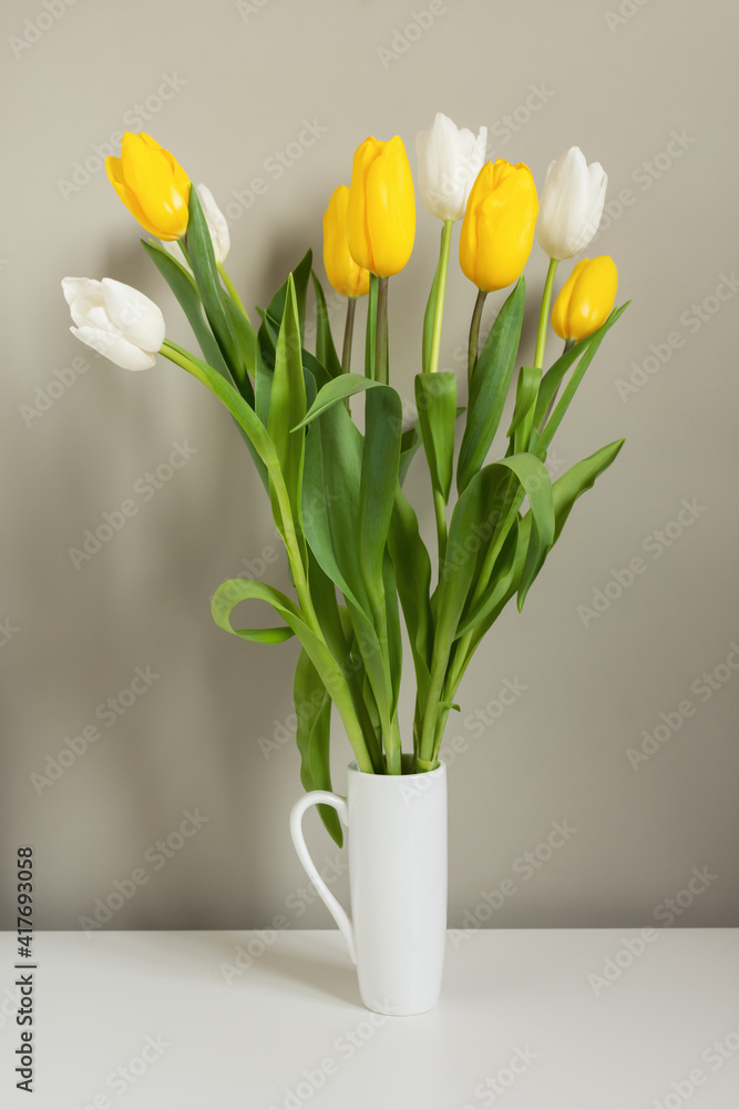 Yellow and white tulips in a white vase on a gray background and a white table. Vertical composition, greeting card, festive bouquet. Mother's Day, International Women's Day, Birthday. Hello, spring. Yellow and white tulips in a white vase on a gray background and a white table. Vertical composition, greeting card, festive bouquet. Mother's Day, International Women's Day, Birthday. Hello, spring.