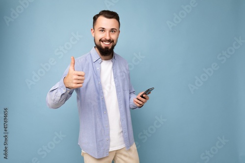 Joyful handsome young brunette unshaven man with beard wearing stylish white t-shirt and blue shirt isolated over blue background with empty space holding in hand and using phone messaging sms looking
