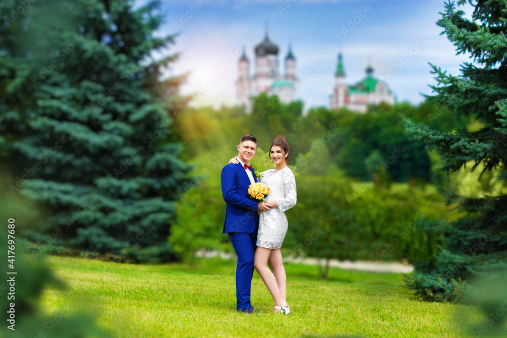 Stylish groom with his beloved beautiful wife, short wedding dress, romantic and cheerful couple in the park against the background of the church