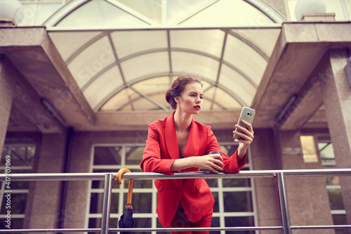 Businesswoman in red costume using mobile phone near office, girl browsing phone, female manager texting smartphone near modern bank building.
