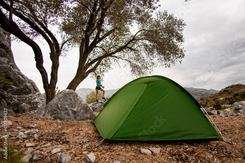 view of tent near rock and trees and woman with trekking sticks nearby.