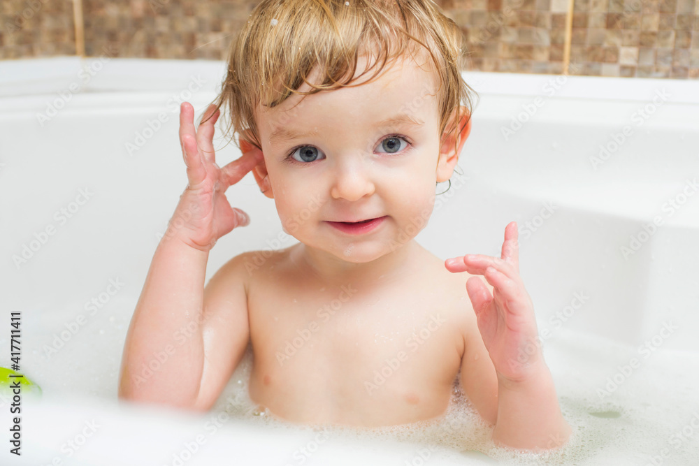 Funny little boy getting a bath in the bathroom. A child bathes in water with soap suds in white bath Funny little boy getting a bath in the bathroom. A child bathes in water with soap suds in white bath
